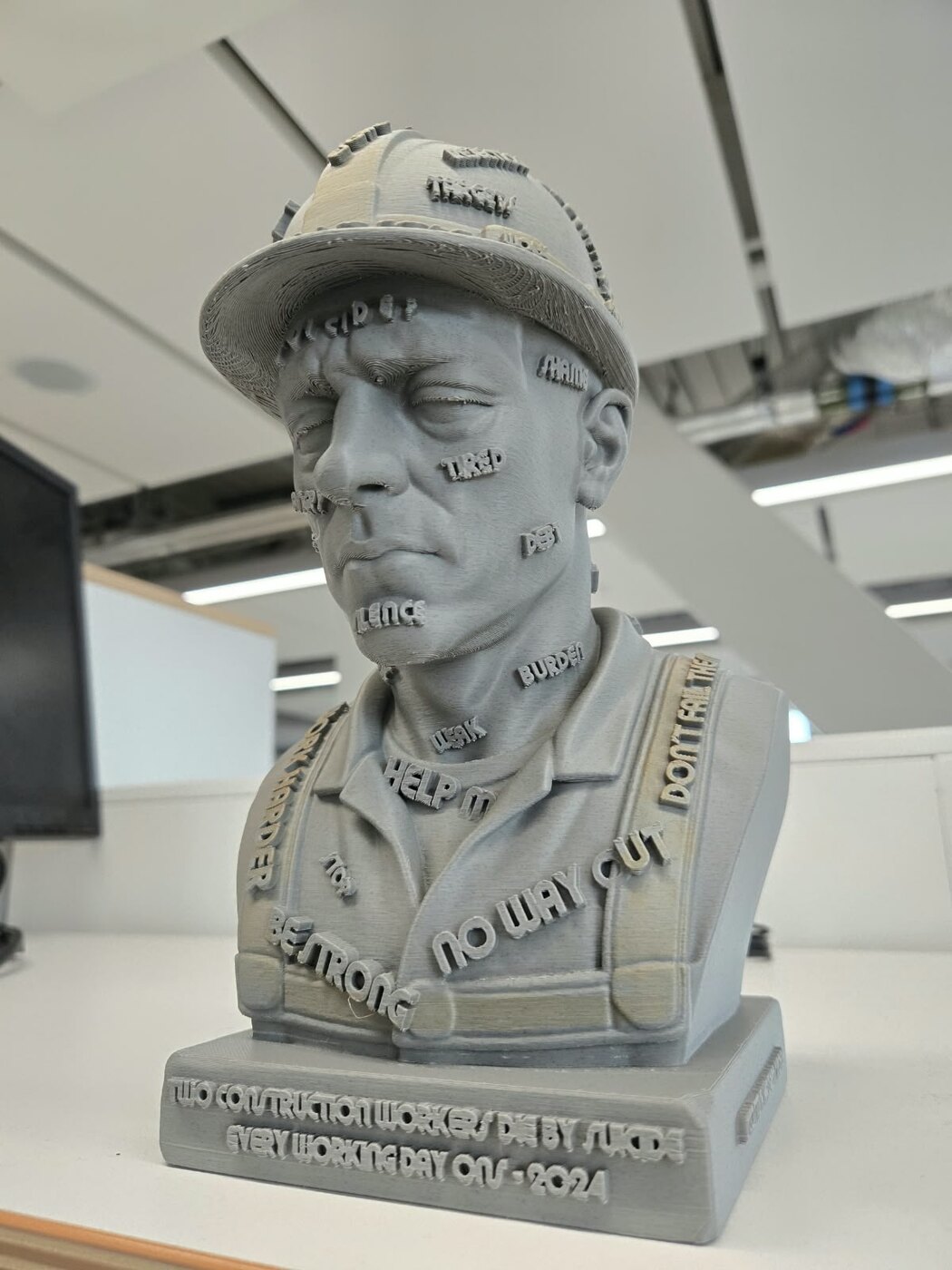 Grey 3D-printed SOLVE Construction Bust viewed from the front, with words describing workplace pressures and distress across the figure