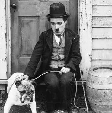 A man wearing a bowler hat and a suit sitting on a wooden step with a bulldog on a leash, in front of a wooden door and a barrel.